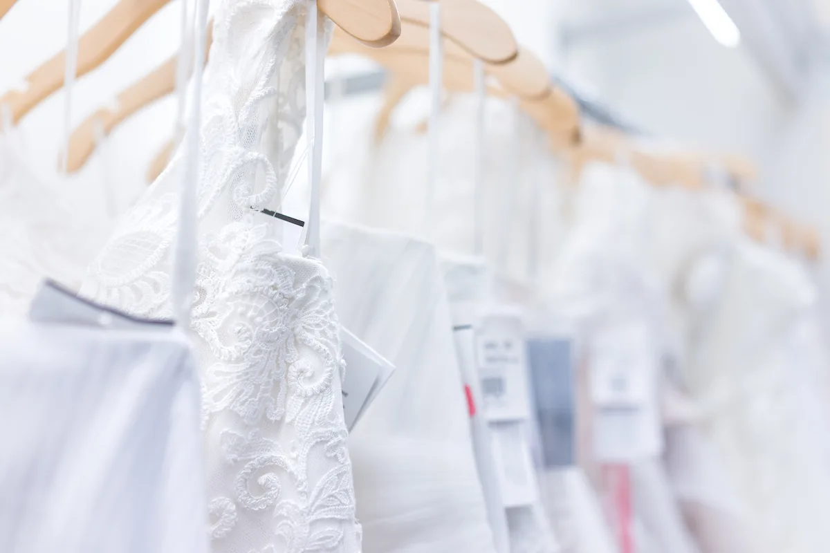 Many wedding dresses with price tags in boutique discount store, white garments hanging on rack hangers row closeup with white lace, tulle, design